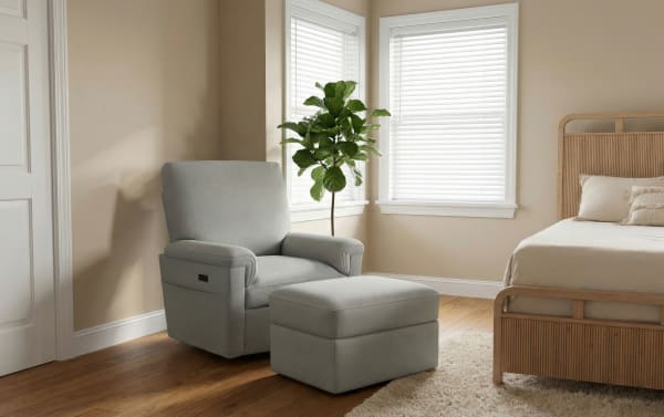 Gray fabric recliner chair with matching ottoman in a beige bedroom, featuring wooden floor, rattan bed frame, and natural light from windows.